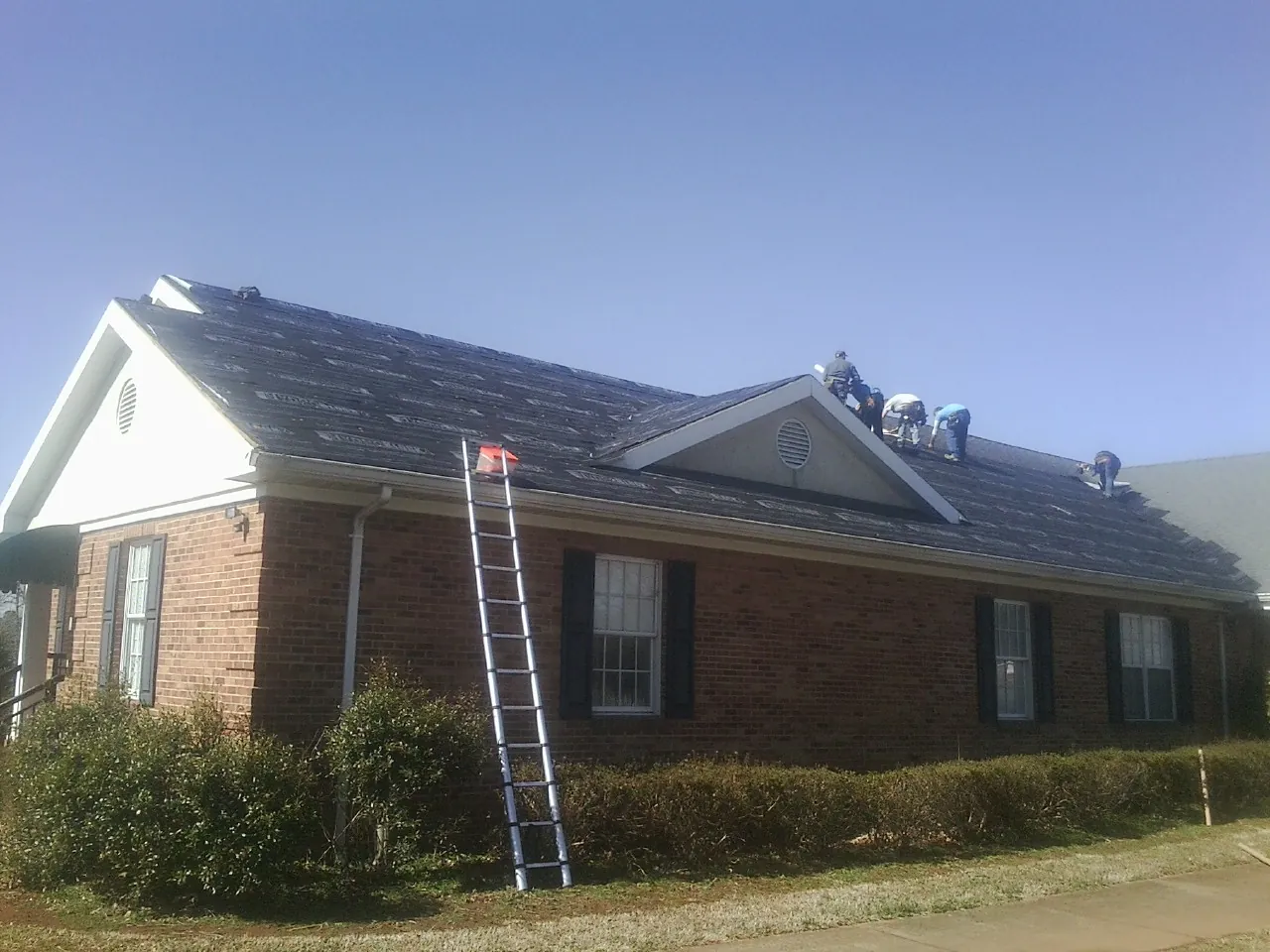 Crew tearing shingles off a brick ranch roof with a ladder set — Emergency Roof Repair work by our crew in Polk City